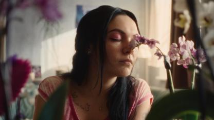 Szene aus dem Film „Vena“: Jenny (gespielt von Emma Nova) mit pinkem Lidschatten und pinkem Shirt sowie langen schwarzen Haaren riecht mit geschlossenen Augen an einer Orchidee.